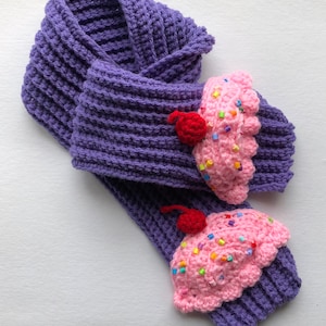 Puede incluir: Bufanda morada de ganchillo con dos dise&ntilde;os de cupcake. Los cupcakes son rosas con chispas de colores y una cereza roja encima. La bufanda est&aacute; hecha de un hilo grueso y texturizado.