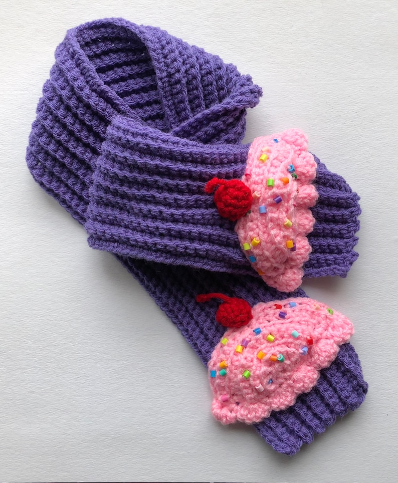 Puede incluir: Bufanda morada de ganchillo con dos dise&ntilde;os de cupcake. Los cupcakes son rosas con chispas de colores y una cereza roja encima. La bufanda est&aacute; hecha de un hilo grueso y texturizado.
