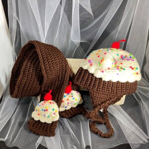 Puede incluir: Un conjunto de crochet hecho a mano con una bufanda marr&oacute;n y un gorro, ambos dise&ntilde;ados para parecerse a cupcakes. El gorro tiene orejeras y una parte superior de glaseado blanco con chispas de colores y una cereza roja. La bufanda tiene extremos de cupcake.