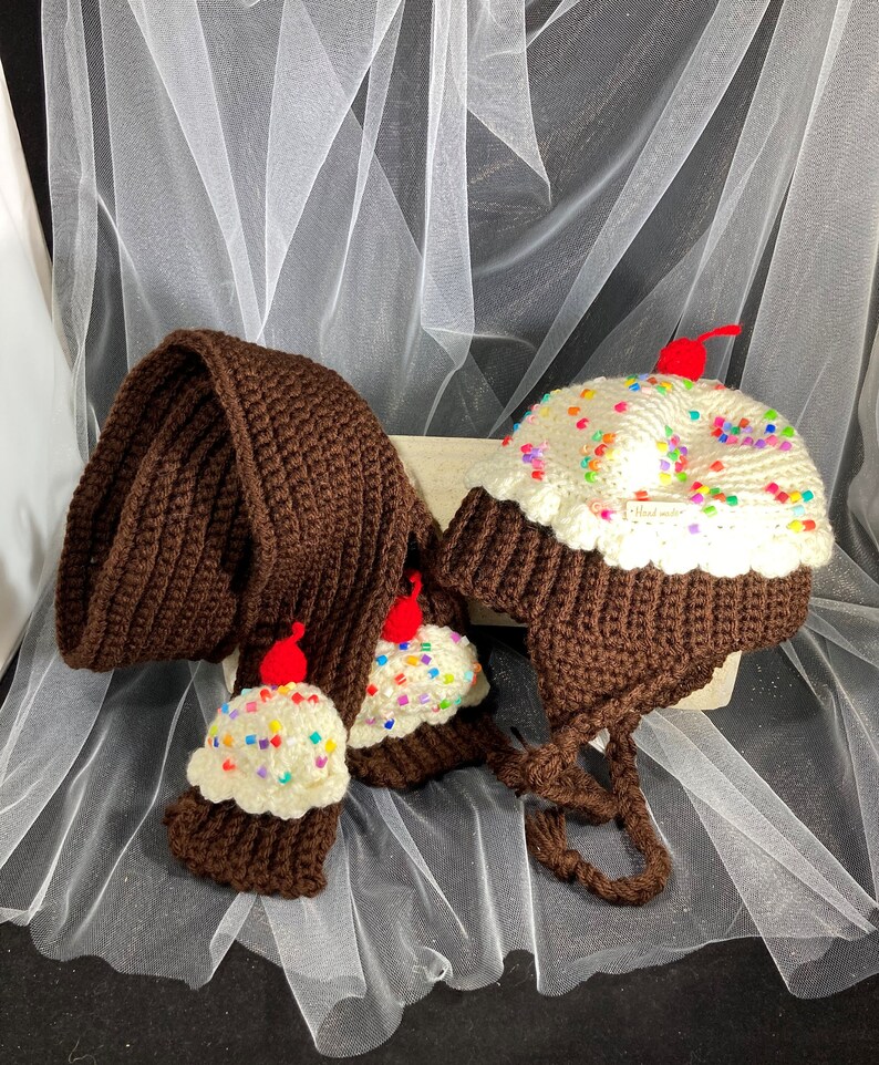 Puede incluir: Un conjunto de crochet hecho a mano con una bufanda marr&oacute;n y un gorro, ambos dise&ntilde;ados para parecerse a cupcakes. El gorro tiene orejeras y una parte superior de glaseado blanco con chispas de colores y una cereza roja. La bufanda tiene extremos de cupcake.