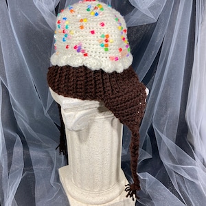 Puede incluir: Un gorro de ganchillo caprichoso con forma de helado. El gorro tiene una base marr&oacute;n, una parte superior blanca cremosa adornada con chispas de colores y una cereza roja. Incluye orejeras y borlas, ofreciendo un aspecto divertido y &uacute;nico.