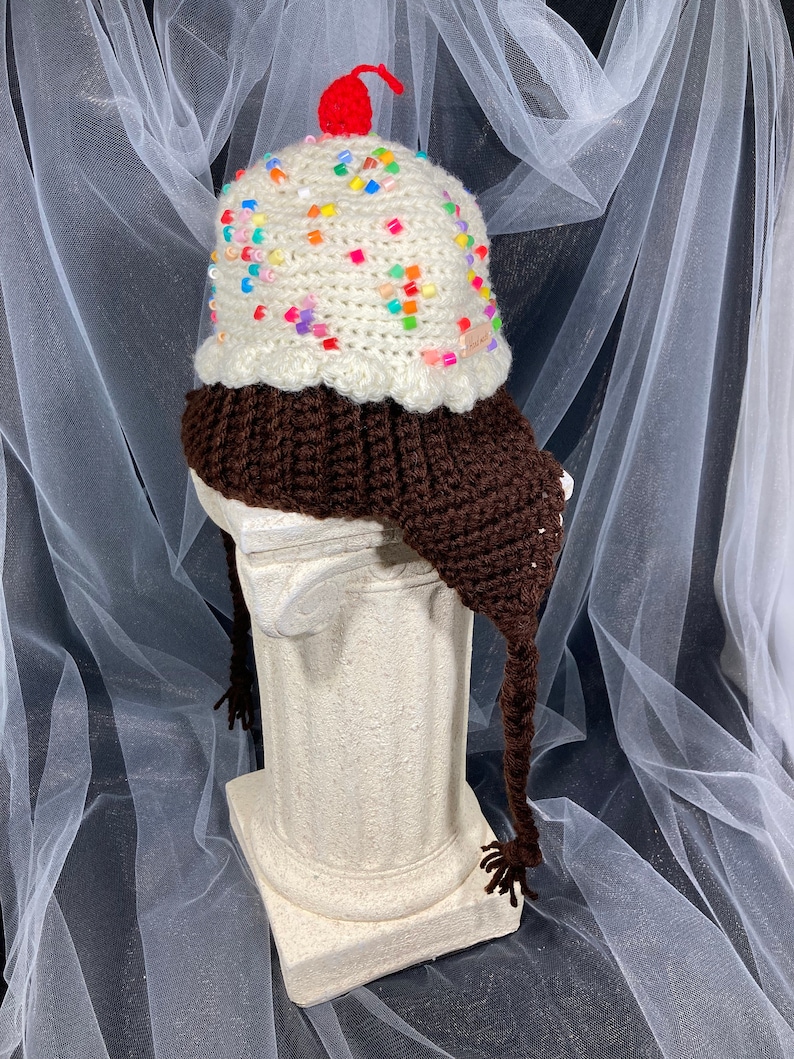 Puede incluir: Un gorro de ganchillo caprichoso con forma de helado. El gorro tiene una base marr&oacute;n, una parte superior blanca cremosa adornada con chispas de colores y una cereza roja. Incluye orejeras y borlas, ofreciendo un aspecto divertido y &uacute;nico.