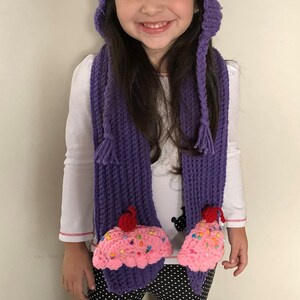 Puede incluir: Un ni&ntilde;o lleva un conjunto de gorro y bufanda de cupcake de ganchillo hechos a mano. El gorro es morado con la parte superior rosa, decorado con chispas de colores y una cereza roja. La bufanda a juego tiene extremos en forma de cupcake. El ni&ntilde;o lleva una camisa blanca y pantalones de lunares blancos y negros.