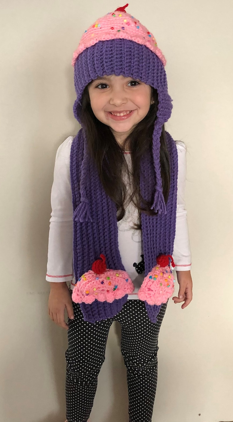 Puede incluir: Un ni&ntilde;o lleva un conjunto de gorro y bufanda de cupcake de ganchillo hechos a mano. El gorro es morado con la parte superior rosa, decorado con chispas de colores y una cereza roja. La bufanda a juego tiene extremos en forma de cupcake. El ni&ntilde;o lleva una camisa blanca y pantalones de lunares blancos y negros.
