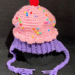 May include: A pink and purple crocheted hat shaped like a cupcake with a red cherry on top. The hat has colourful sprinkles and a long purple tassel.