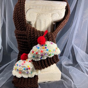 Puede incluir: Una bufanda marr&oacute;n tejida a ganchillo con dos adornos en forma de cupcake. Los cupcakes tienen glaseado blanco con chispas de colores y una cereza roja. La bufanda est&aacute; envuelta alrededor de una estatua blanca.
