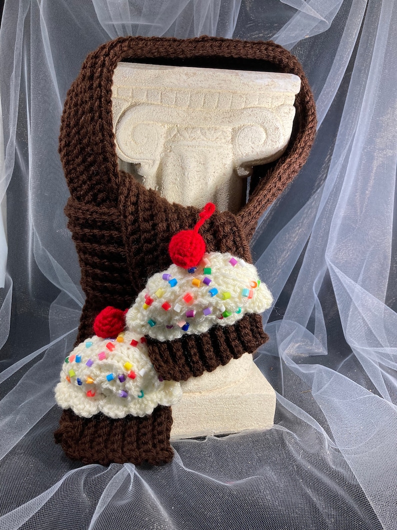 Puede incluir: Una bufanda marr&oacute;n tejida a ganchillo con dos adornos en forma de cupcake. Los cupcakes tienen glaseado blanco con chispas de colores y una cereza roja. La bufanda est&aacute; envuelta alrededor de una estatua blanca.