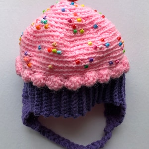 Puede incluir: Un gorro de cupcake de ganchillo hecho a mano con la parte superior rosa, la base morada y una cereza roja. La cobertura rosa est&aacute; decorada con chispas de colores. El gorro tiene lazos trenzados.