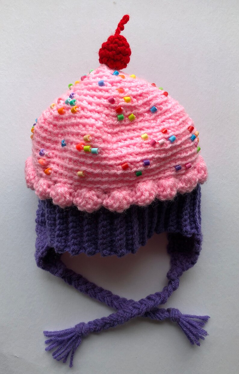 Puede incluir: Un gorro de cupcake de ganchillo hecho a mano con la parte superior rosa, la base morada y una cereza roja. La cobertura rosa est&aacute; decorada con chispas de colores. El gorro tiene lazos trenzados.