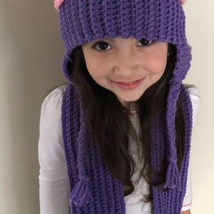 Puede incluir: Un conjunto de gorro y bufanda de cupcake hechos a mano con ganchillo. El gorro es morado con la parte superior rosa, decorado con chispas de colores y una cereza roja. La bufanda a juego tiene detalles de cupcake.