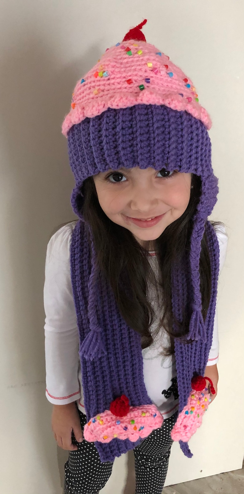 Puede incluir: Un conjunto de gorro y bufanda de cupcake hechos a mano con ganchillo. El gorro es morado con la parte superior rosa, decorado con chispas de colores y una cereza roja. La bufanda a juego tiene detalles de cupcake.