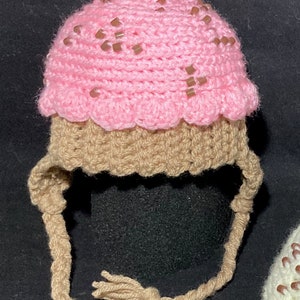 May include: A pink and brown crocheted hat shaped like an ice cream cone with a red cherry on top. The hat has long brown ties.