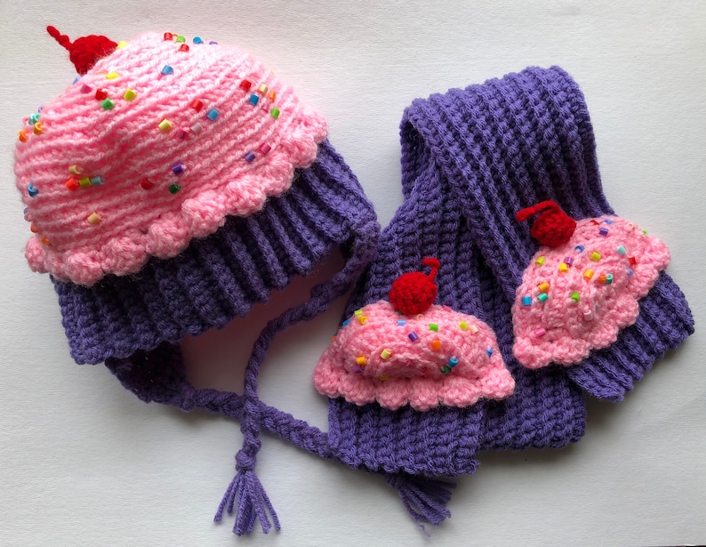 Puede incluir: Un conjunto de gorro y bufanda de punto con tem&aacute;tica de cupcake. El gorro est&aacute; dise&ntilde;ado para parecerse a un cupcake con glaseado rosa, chispas y una cereza roja encima. La bufanda a juego tiene aplicaciones de cupcake.
