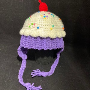May include: Purple and white crocheted hat shaped like a cupcake with colourful sprinkles and a red cherry on top. The hat has long ties that can be tied under the chin.