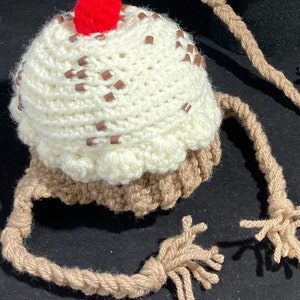 May include: A crocheted ice cream cone hat with a red cherry on top and brown strings. The hat is white with brown sprinkles.