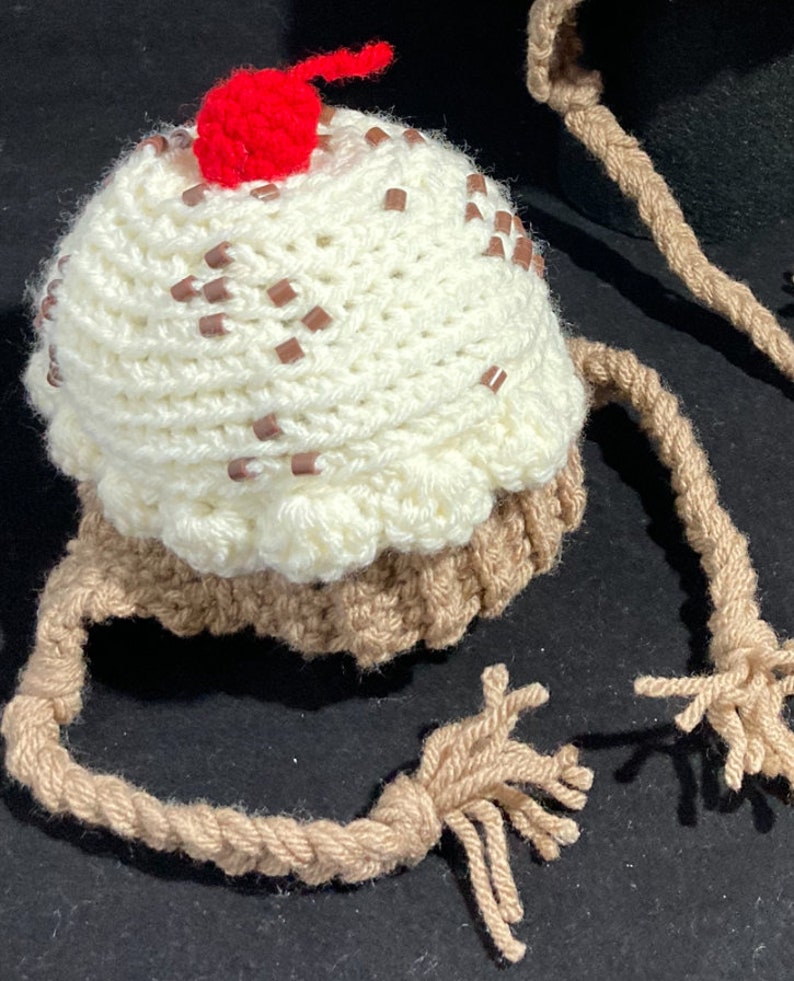 May include: A crocheted ice cream cone hat with a red cherry on top and brown strings. The hat is white with brown sprinkles.