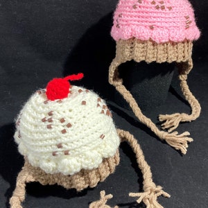 May include: Two crocheted cupcake hats with brown yarn ties. One hat is pink with brown sprinkles and the other is white with brown sprinkles.