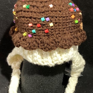 May include: A brown crocheted hat shaped like an ice cream cone with colourful sprinkles and a red cherry on top. The hat has long white crocheted ear flaps.