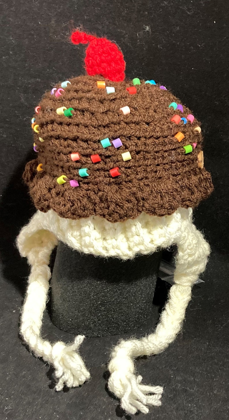 May include: A brown crocheted hat shaped like an ice cream cone with colourful sprinkles and a red cherry on top. The hat has long white crocheted ear flaps.