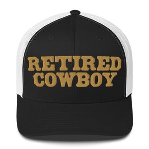 Op de afbeelding: Zwarte en witte truckercap met een zwart voorpaneel en een wit mesh achterpaneel. Het voorpaneel heeft de tekst "RETIRED COWBOY" geborduurd in goud.