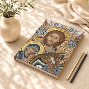 Può includere: Un taccuino rettangolare con una colorata illustrazione in stile mosaico sulla copertina. L'opera raffigura una figura centrale con un'aureola dorata, una donna con un bambino e un angelo, il tutto realizzato con piccole e complesse tessere.