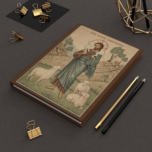 May include: A hardback journal featuring a brown cover and an illustration of "The Good Shepherd." The image shows a man with a lamb, surrounded by sheep, with text above. Gold and black pencils and paper clips are also visible.