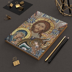 May include: A hardcover journal with a mosaic-style religious image on the cover. The artwork features a depiction of Jesus, Mary, and an angel, with a gold-colored border. Gold binder clips and pencils are also visible.