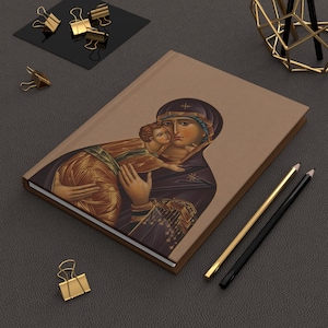 Puede incluir: Un diario de tapa dura con un icono religioso de una madre y un niño. La portada es de color marrón claro con la imagen en colores ricos. Lápices dorados y negros y clips dorados están colocados alrededor del diario.