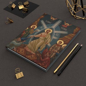Puede incluir: Un diario de tapa dura con una ilustración de icono religioso. La portada representa una figura central con un halo, rodeada de otras figuras y ángeles. La paleta de colores incluye azules, marrones y rojos. También se ven lápices dorados y negros y clips dorados.
