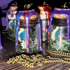 May include: Small glass pendant bottles filled with greenery, blue and white orbs, and star-shaped charms. The corked tops are sealed with dripping purple, red, and blue wax. A gold-colored chain is in the foreground.
