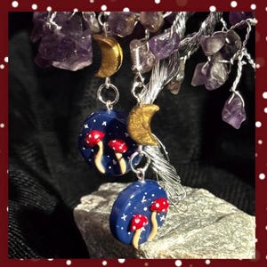 May include: A pair of handmade earrings featuring a crescent moon charm and a round blue charm with white stars and red and white mushrooms.