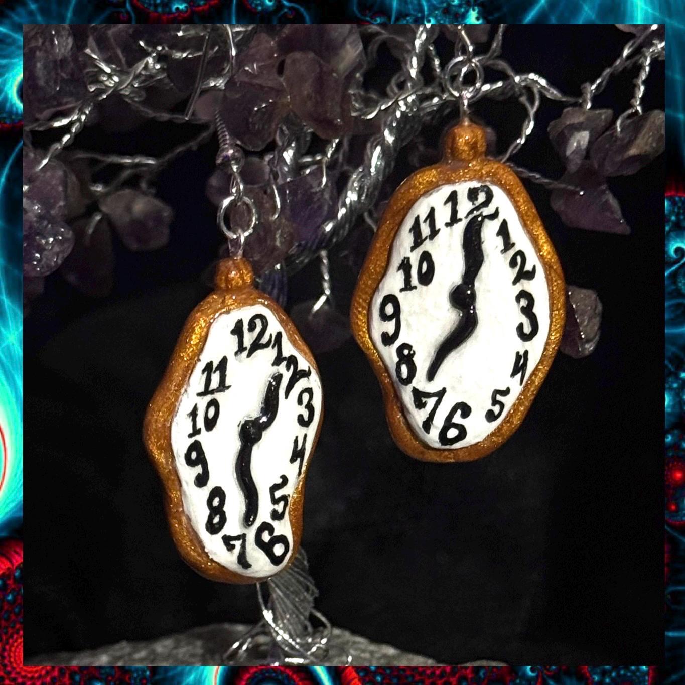 May include: A pair of gold and white earrings shaped like melting clocks. The clocks have black hands and black numbers. The earrings are hanging from silver hooks.