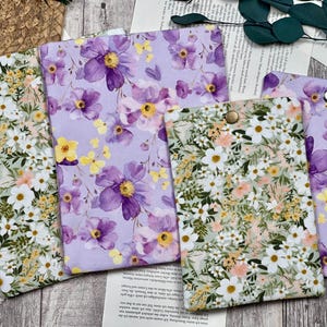Può includere: Una collezione di custodie in tessuto con motivi floreali. Le custodie presentano disegni con fiori viola, accenti gialli e fiori bianchi con fogliame verde. Una custodia ha una chiusura a bottone. Le custodie sono disposte su una superficie di legno.