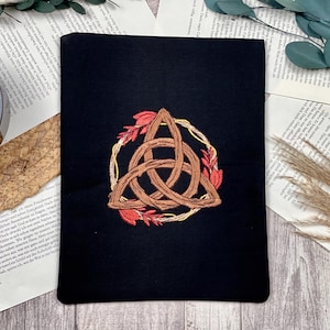 May include: Black book cover featuring a brown Celtic knot design embroidered within a gold wreath, accented with red leaves. The cover is rectangular and made of a dark fabric. The design is centered on the cover.