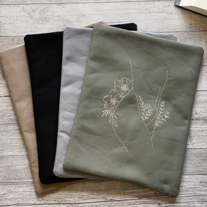 Op de afbeelding: Vier stoffen zakjes in verschillende kleuren, waaronder beige, zwart, grijs en groen. Het groene zakje heeft een wit geborduurd bloemenmotief.