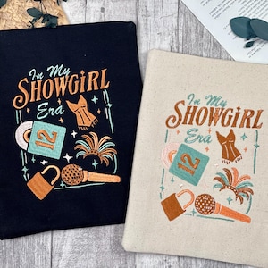 Puede incluir: Dos bolsas de mano, una negra y otra beige, con el texto bordado "In My Showgirl Era". Cada bolsa presenta un corsé, un micrófono, un disco y un diseño de cerradura en naranja, turquesa y dorado.