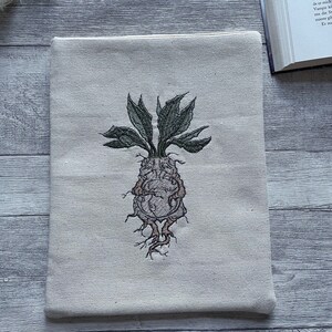 Op de afbeelding: Een beige canvas laptophoes met een geborduurd beeld van een groene plant met een bruin wortelstelsel.