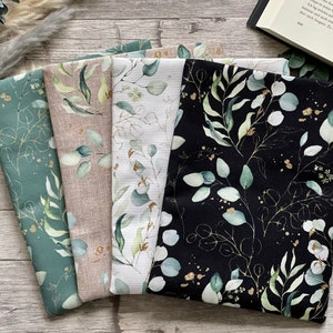 Könnte beinhalten: Eine Sammlung von Stoffstücken mit botanischen Designs. Die Stoffe zeigen Eukalyptusblätter in Grün-, Gold- und Weißtönen auf Hintergründen in Türkis, Beige und Schwarz. Die Stoffe sind gefaltet und auf einer Holzoberfläche angeordnet.
