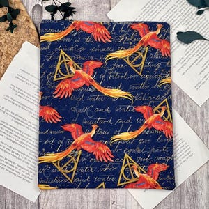 May include: A navy blue book cover featuring a repeating pattern of red and yellow phoenixes and gold Deathly Hallows symbols. The cover also has handwritten script in gold. The overall theme is fantasy.