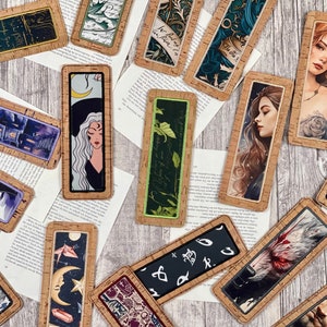 Pode incluir: Uma coleção de marcadores de página com suporte de cortiça com vários designs, incluindo um gato preto, uma bruxa, um lobo e um padrão floral. Os marcadores de página são todos retangulares e têm um suporte de cortiça castanho.