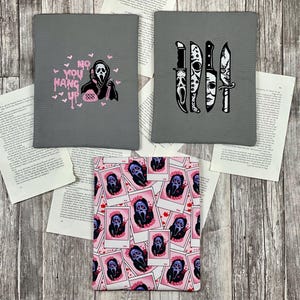 Könnte beinhalten: Drei verschiedene Designs für einen Stoffbuchbezug. Das erste Design ist grau mit einem rosa Geist, der ein Telefon hält, und dem Text "No You Hang Up". Das zweite Design ist grau mit vier schwarzen und weißen Messern. Das dritte Design ist rosa mit einem sich wiederholenden Muster eines Geistes in einem weißen Rahmen.