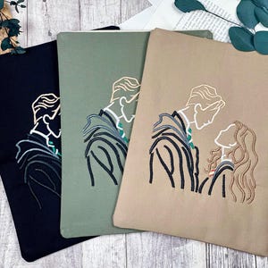 May include: Three fabric pouches in black, sage green, and tan, each embroidered with a line-art illustration of a couple. The design features a man and woman in profile, with the man wearing a striped jacket and the woman with flowing hair.