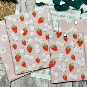 Könnte beinhalten: Rosa Stoffartikel mit einem Muster aus roten Erdbeeren und weißen Blumen. Die Artikel umfassen einen Buchumschlag, einen kleinen Beutel mit Druckknopfverschluss und andere rechteckige Teile. Der Hintergrund umfasst einen geflochtenen Korb und Grünzeug.