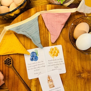 Pode incluir: Uma mesa de madeira exibe ovos em tigelas de vidro, uma faixa de tecido colorida e cartões inspiradores. Os cartões apresentam ilustrações de mirtilos, um favo de mel e uma lanterna, com versículos bíblicos. Também há um pegador de mel de madeira.