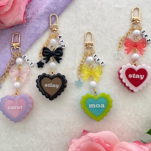 May include: Four keychains with gold-colored clasps, each featuring a different design. They include heart-shaped pendants with text, bows, star charms, and beaded chains. The keychains have the words "carat", "atiny", "moa", and "stay".