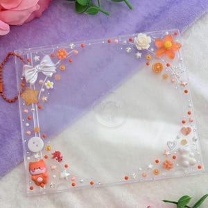 May include: Clear plastic CD case decorated with various embellishments. The case features a white bow, orange flowers, stars, and a small orange fox. The edges are lined with orange and white beads. A small chain is attached to the side.