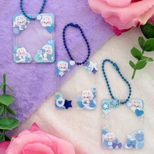 May include: Three clear acrylic keychains with blue ball chains. Each keychain features a rectangular frame with rounded corners, decorated with cartoon bunnies and blue accents. The keychains are displayed on a purple and white surface with pink roses.