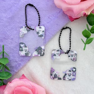 May include: Two clear acrylic keychains with black ball chains. Each keychain features a square frame with a purple and white cat design, a haunted house, and hearts. The keychains are displayed on a purple and white background with pink roses.