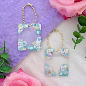 May include: Two clear, rectangular keychains with a gold-colored chain. Each keychain features a cartoon bunny design with blue, green, and white accents. The word "Cute" is printed on one keychain. The background is a mix of purple and white.