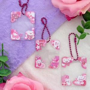 May include: Three clear acrylic keychains with pink and white bear and star designs. Each keychain has a pink ball chain. One keychain has the text "Enjoy June". The background includes pink roses and a purple cloth.